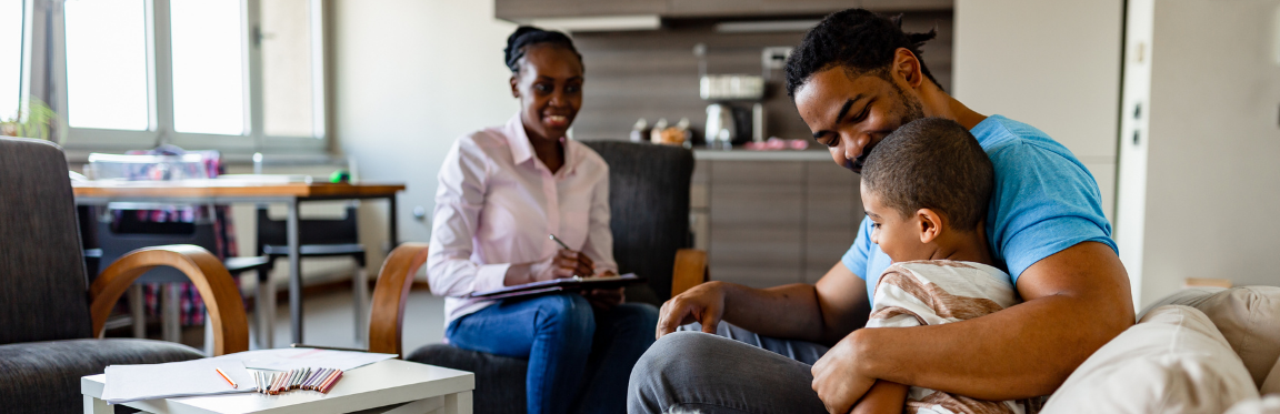 A practitioner sits across from a father and his child in a living room. The practitioner smiles and takes notes.