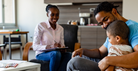 A practitioner sits across from a father and his child in a living room. The practitioner smiles and takes notes.