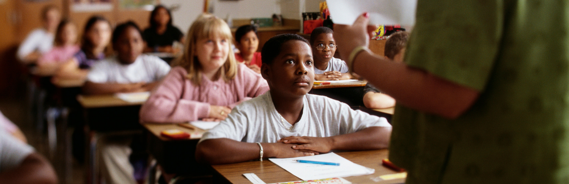 A classroom of students sit at their desk with a pencil and paper in front of them, looking up at their teacher.