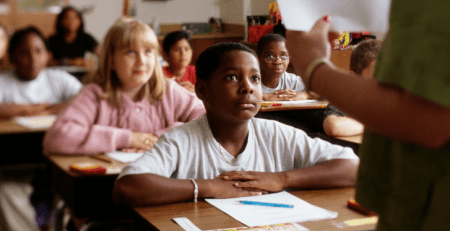 A classroom of students sit at their desk with a pencil and paper in front of them, looking up at their teacher.