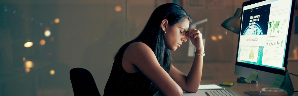 A woman sits at her computer and looks stressed. She is holding her forehead with her eyes closed.