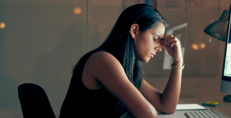 A woman sits at her computer and looks stressed. She is holding her forehead with her eyes closed.