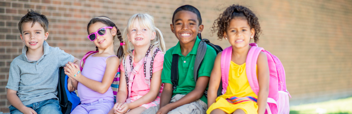 A group of children sit next to each other with their backpacks on and smile.