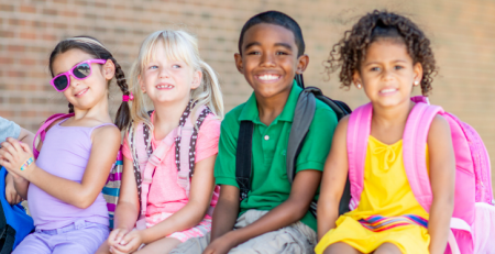 A group of children sit next to each other with their backpacks on and smile.