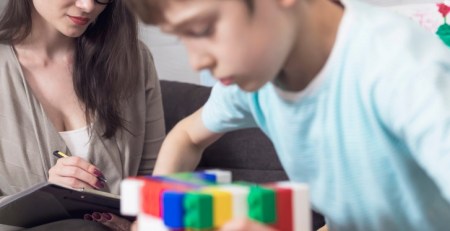 A school psychologist sits with a child and takes notes as the child plays with Lego.