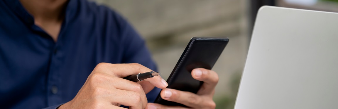 A person works on their laptop, holding a pen and their phone.