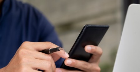 A person works on their laptop, holding a pen and their phone.