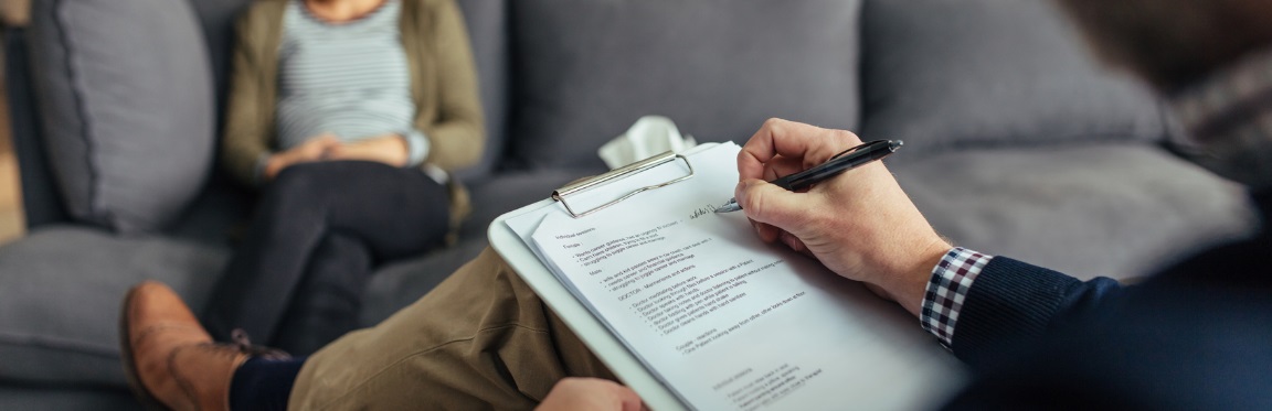 A practitioner sits across from their client. They take notes on their clipboard.