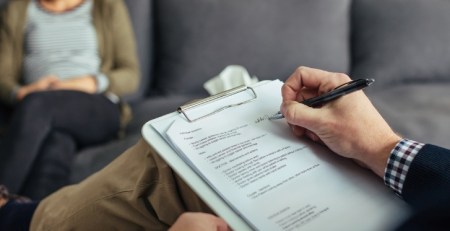 A practitioner sits across from their client. They take notes on their clipboard.