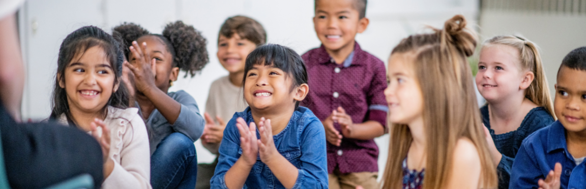 A group of children sit together facing their teacher. They smile and clap their hands.