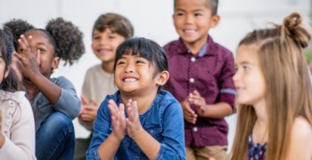 A group of children sit together facing their teacher. They smile and clap their hands.