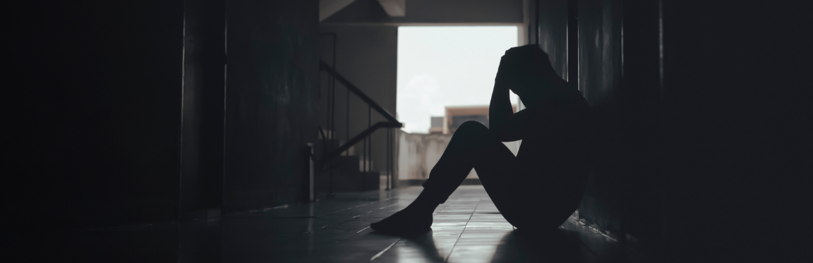 A person sits on the floor of a dark hallway. They hold their head and face the floor.