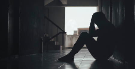 A person sits on the floor of a dark hallway. They hold their head and face the floor.