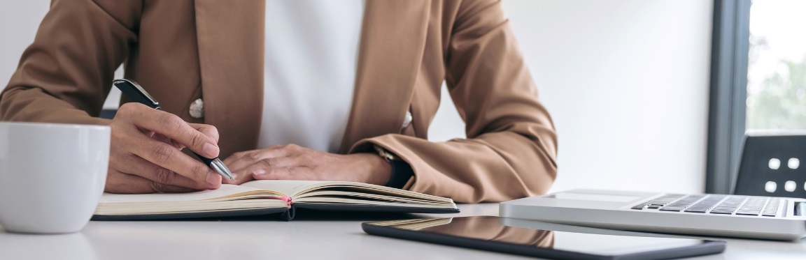 A person sitting at their desk, writing in their notebook, with their laptop and tablet in front of them.