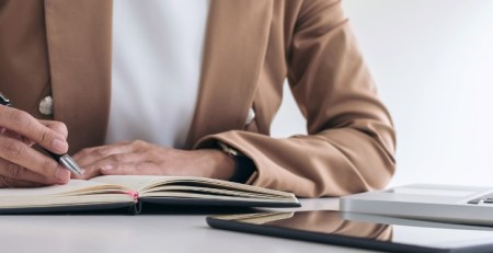 A person sitting at their desk, writing in their notebook, with their laptop and tablet in front of them.