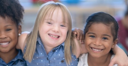 Three young girls smile and pose for a photo. The girl in the middle has her arms around the two girls beside her.