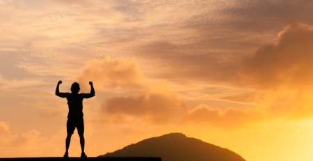 A person stands firmly on top of a high structure with their fists up in the air. Behind them is a mountain top and a glowing sunset.