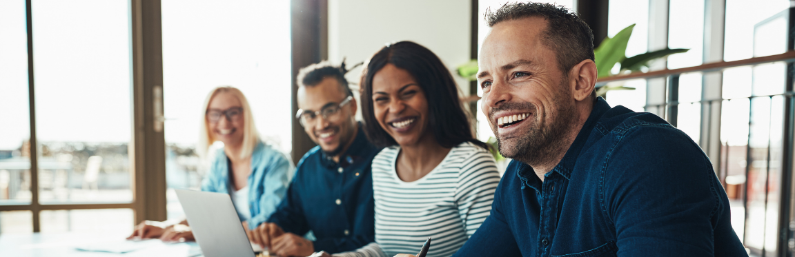 Four colleagues sit next to each other at a conference table and smile.