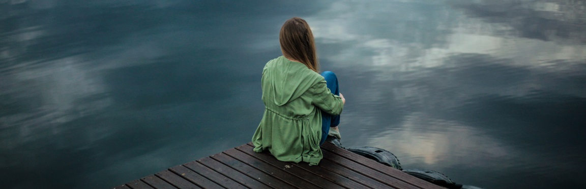 A girl sits on the edge of a dock. She faces a body of water.