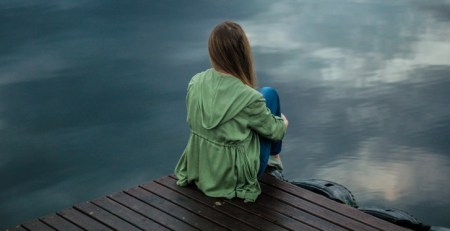 A girl sits on the edge of a dock. She faces a body of water.