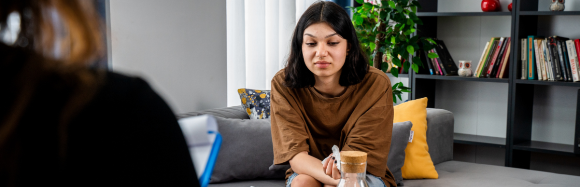 A woman sits on a couch across from a practitioner. The women looks down and appears distressed.