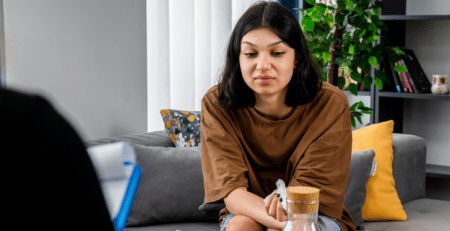 A woman sits on a couch across from a practitioner. The women looks down and appears distressed.