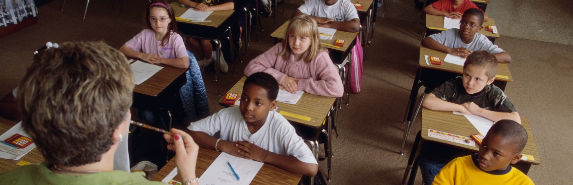 A classroom of students sit at their desk, attentively listening to their teacher.