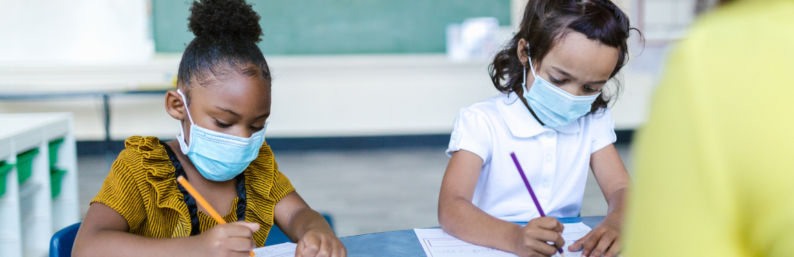 Two young girls sit at their desk in a classroom with their masks on. They concentrate on their work.