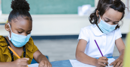 Two young girls sit at their desk in a classroom with their masks on. They concentrate on their work.