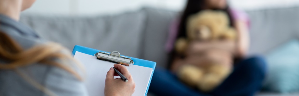 A practitioner holds up a clipboard. In the background, a young girl sits on a couch, holding a teddy bear.