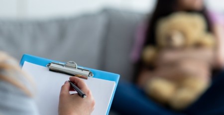 A practitioner holds up a clipboard. In the background, a young girl sits on a couch, holding a teddy bear.