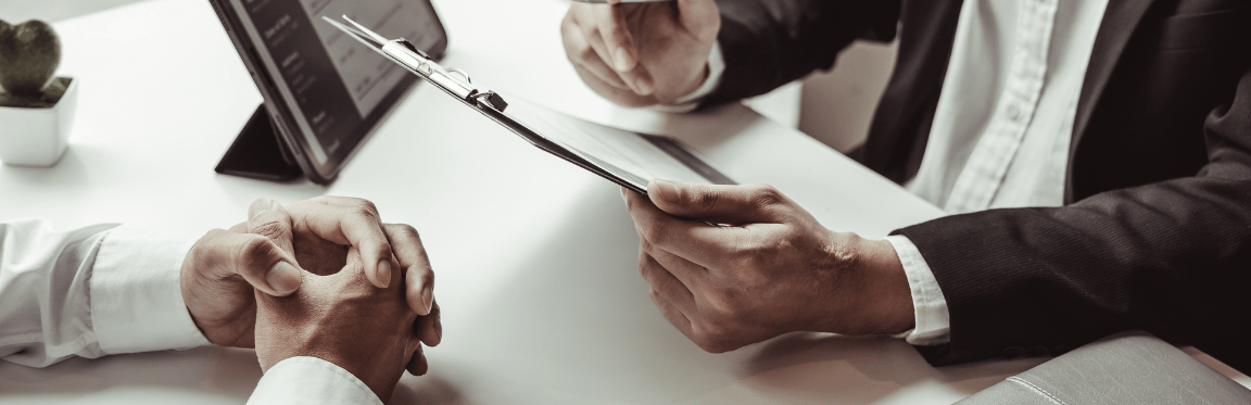 Two professionals sit at a table. One person holds up a clipboard, going over information.