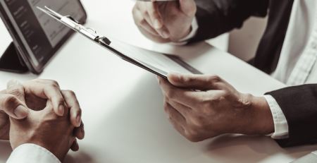 Two professionals sit at a table. One person holds up a clipboard, going over information.