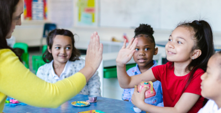 A young student sits at a table with her peers. She is high-fiving her teacher.