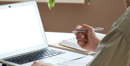 A person sits in front of a laptop screen and writes in their notebook.