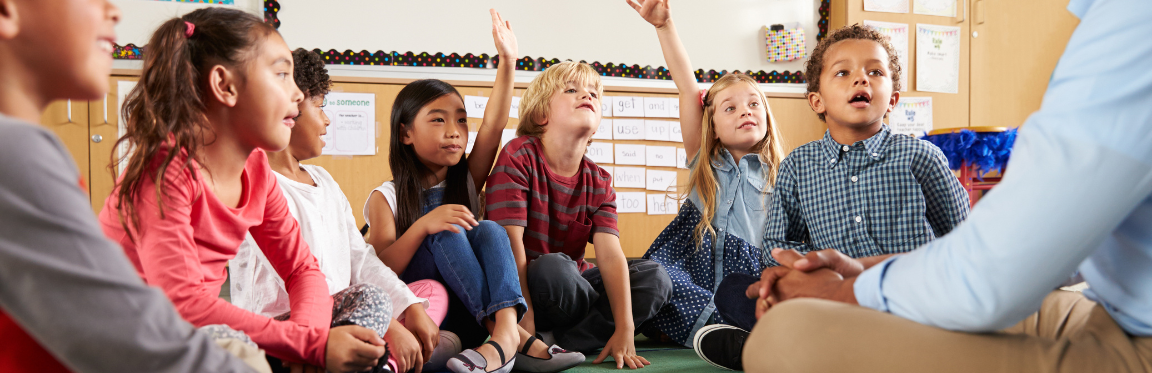 A group of children sit on the floor in their classroom next to their teacher. Two of the students raise their hand.