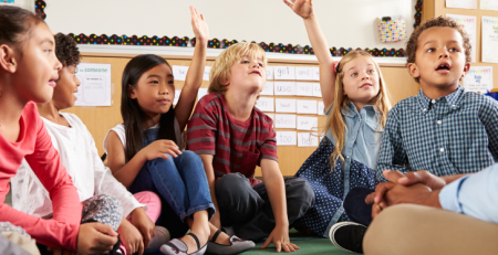 A group of children sit on the floor in their classroom next to their teacher. Two of the students raise their hand.