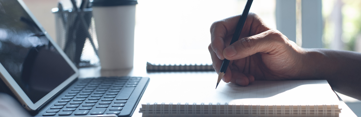 A person's hand writing in a notebook in front of a tablet device.