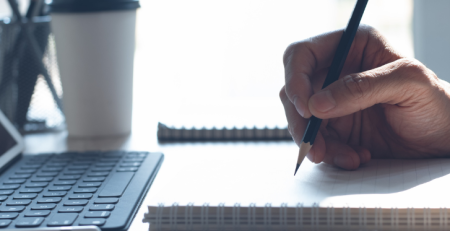 A person's hand writing in a notebook in front of a tablet device.