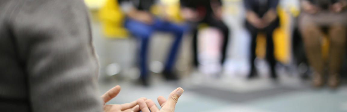 A meeting being held with five people. One person is in focus, talking to the rest of the group, while the other four people are blurred in the background.