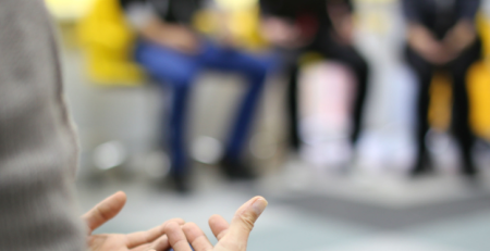 A meeting being held with five people. One person is in focus, talking to the rest of the group, while the other four people are blurred in the background.