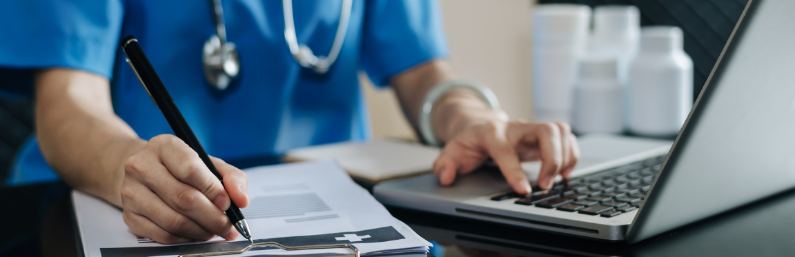 A doctor sits at their desk, working on their laptop, and takes notes on a clipboard.