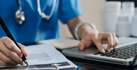 A doctor sits at their desk, working on their laptop, and takes notes on a clipboard.