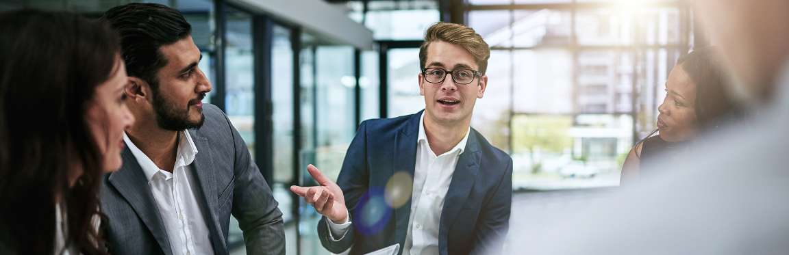 A group of professionals sit in a meeting. One person leads the conversation and the others listen.