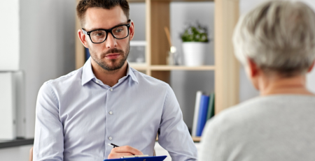 A practitioner is sitting down with their patient, attentively listening and writing down information on a clipboard.