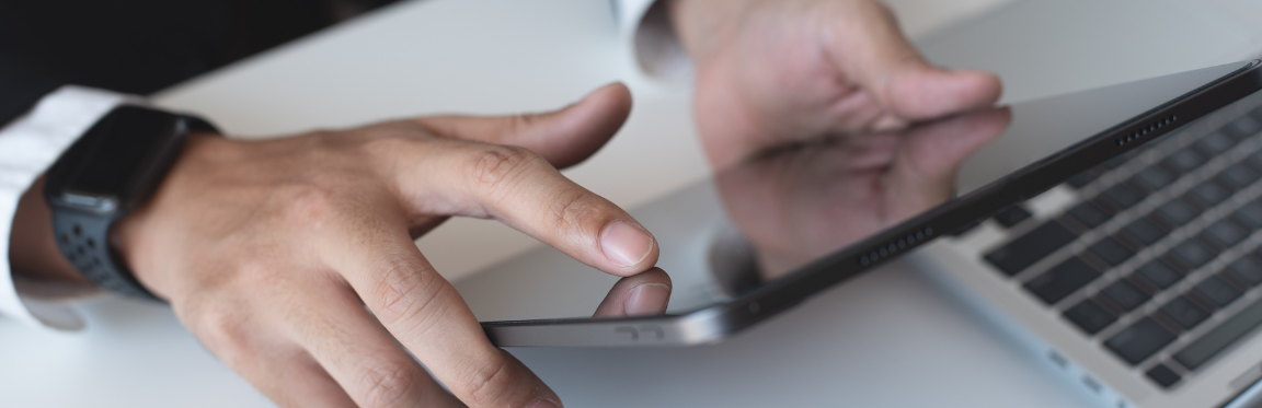 A person’s hands holding up an iPad, wearing an Apple Watch, with a laptop open in front of them.