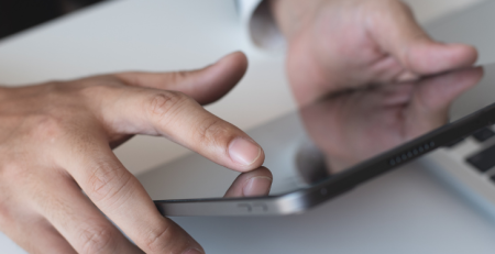 A person’s hands holding up an iPad, wearing an Apple Watch, with a laptop open in front of them.