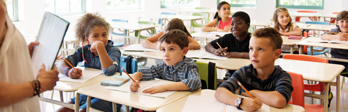 A classroom of children at their desks, listening to their teacher and writing in their notebooks.