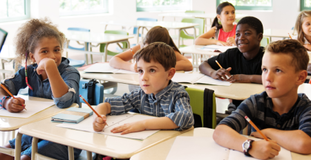 A classroom of children at their desks, listening to their teacher and writing in their notebooks.