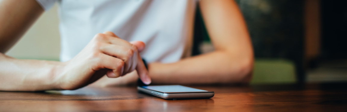 A person sitting at a table, resting their forearms, and viewing their phone.